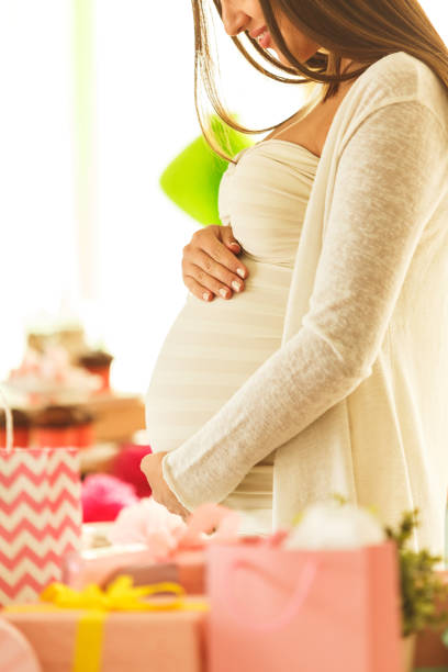 Pregnant woman at baby shower holding her pregnant belly, standing next to stack of presents.